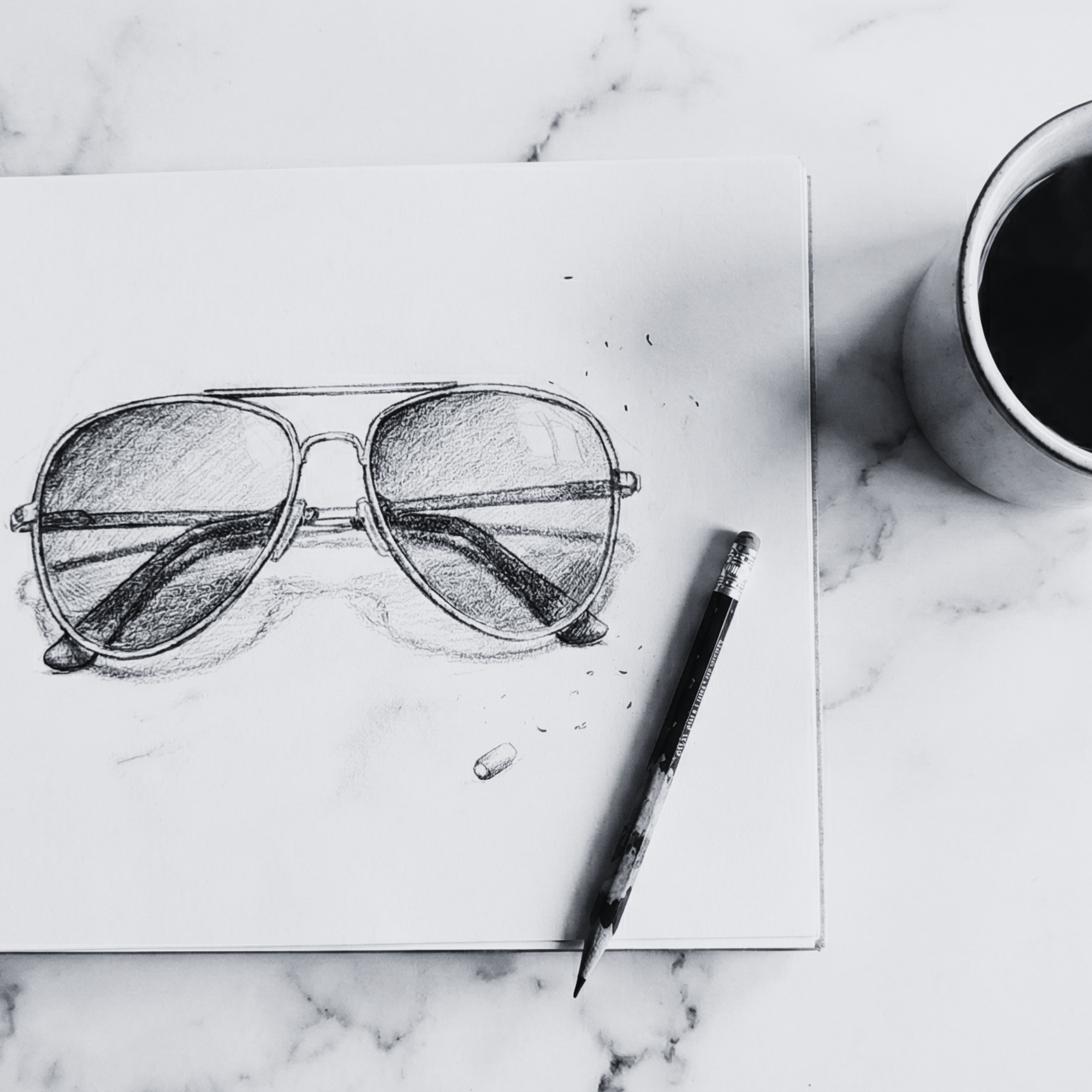 Pencil sketch of sunglasses on a piece of paper with a cup of coffee on a marble surface.