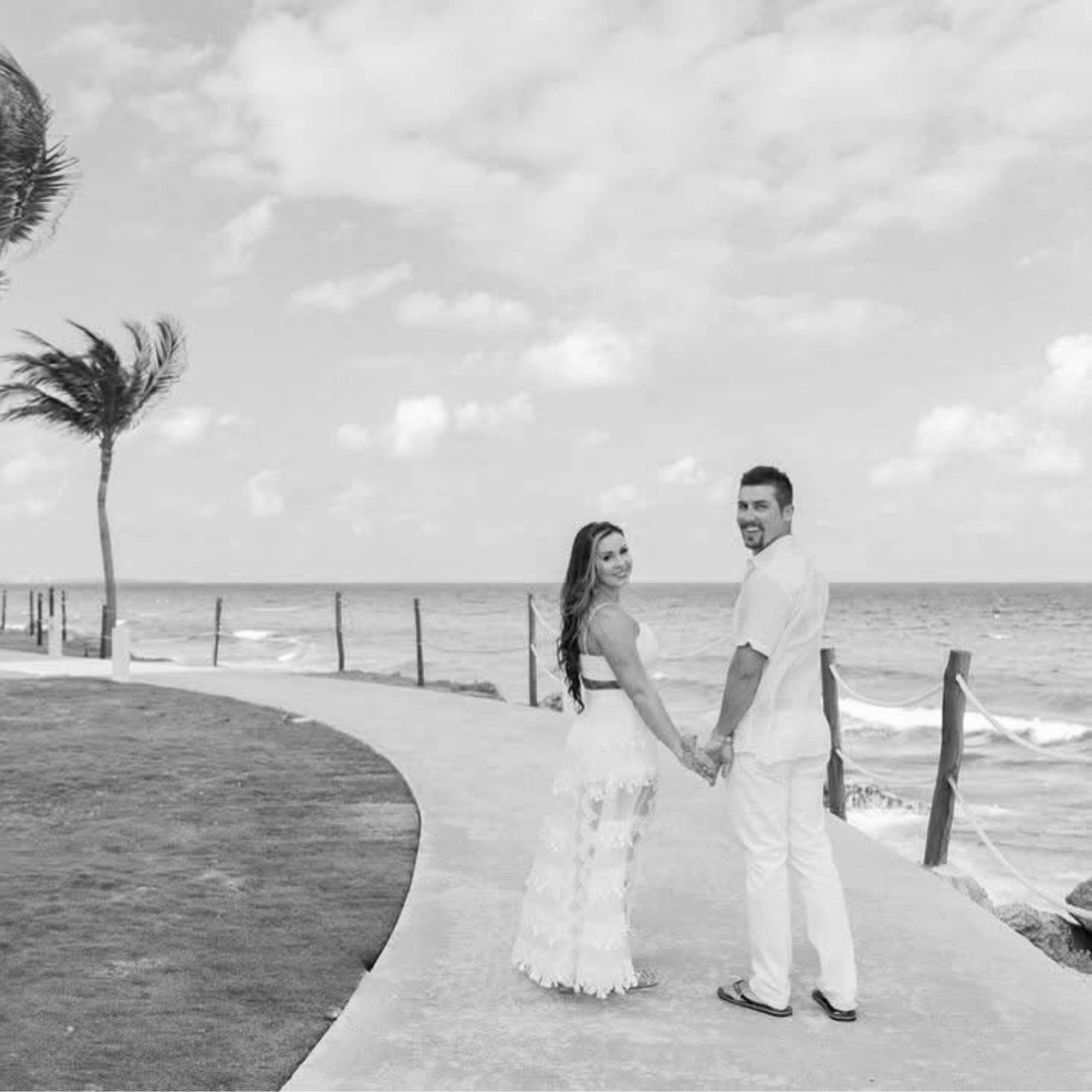 Black and white photo of a couple standing on a beach path with palm trees and ocean in the background.