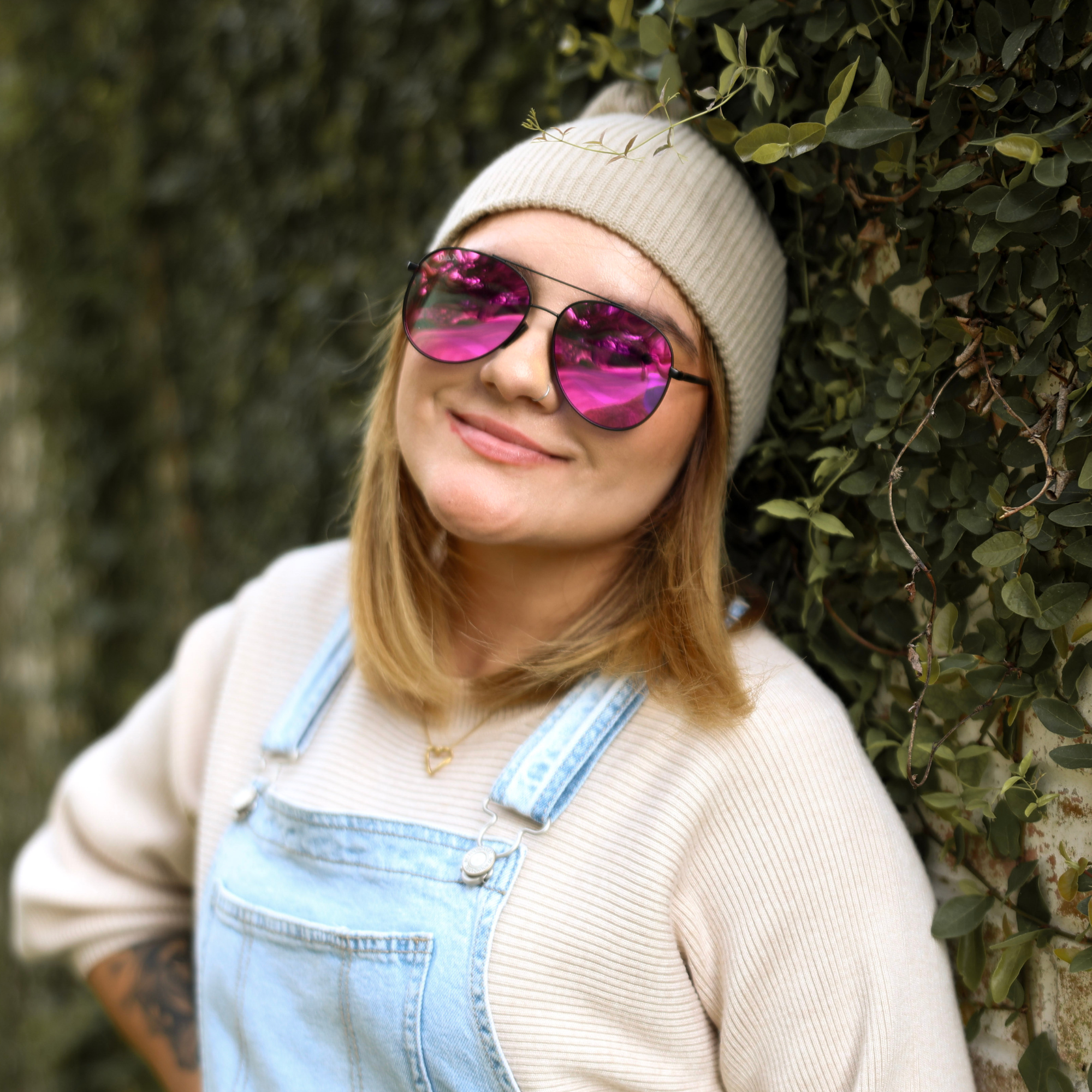 Person wearing pink sunglasses, beige knit hat, and denim overalls standing against a green leafy background.