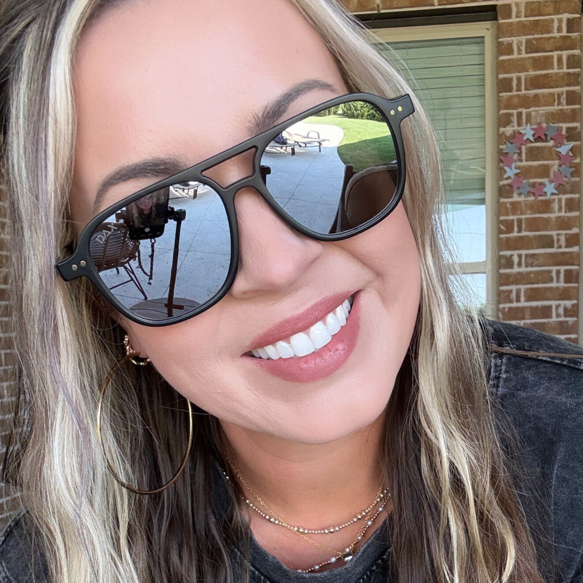 A close-up lifestyle shot of a woman wearing black aviator-style sunglasses with dark mirrored lenses, set against a background of a brick wall and window.