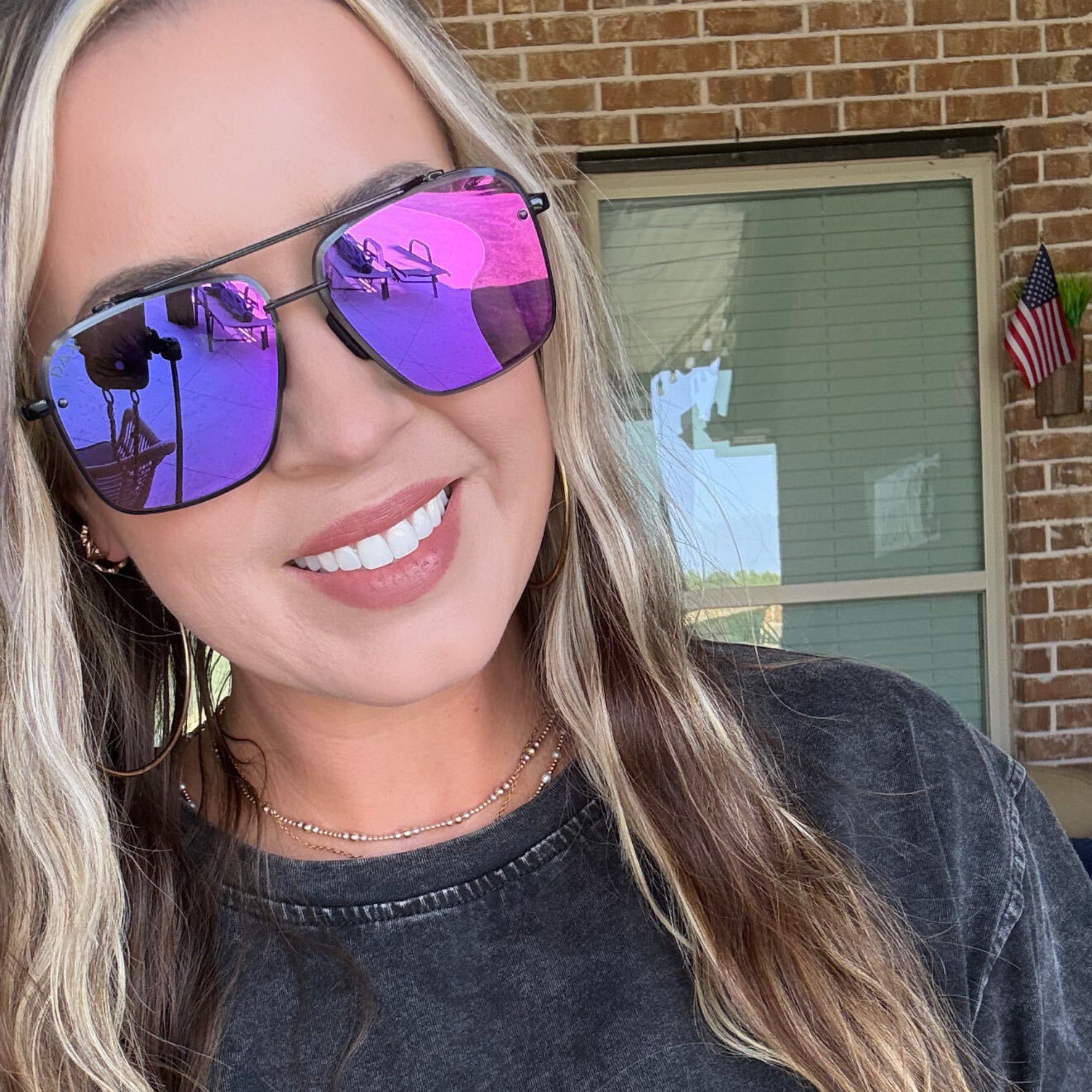 A close-up of a woman wearing square aviator-style sunglasses with vibrant purple mirrored lenses, standing outdoors in front of a brick wall and window.