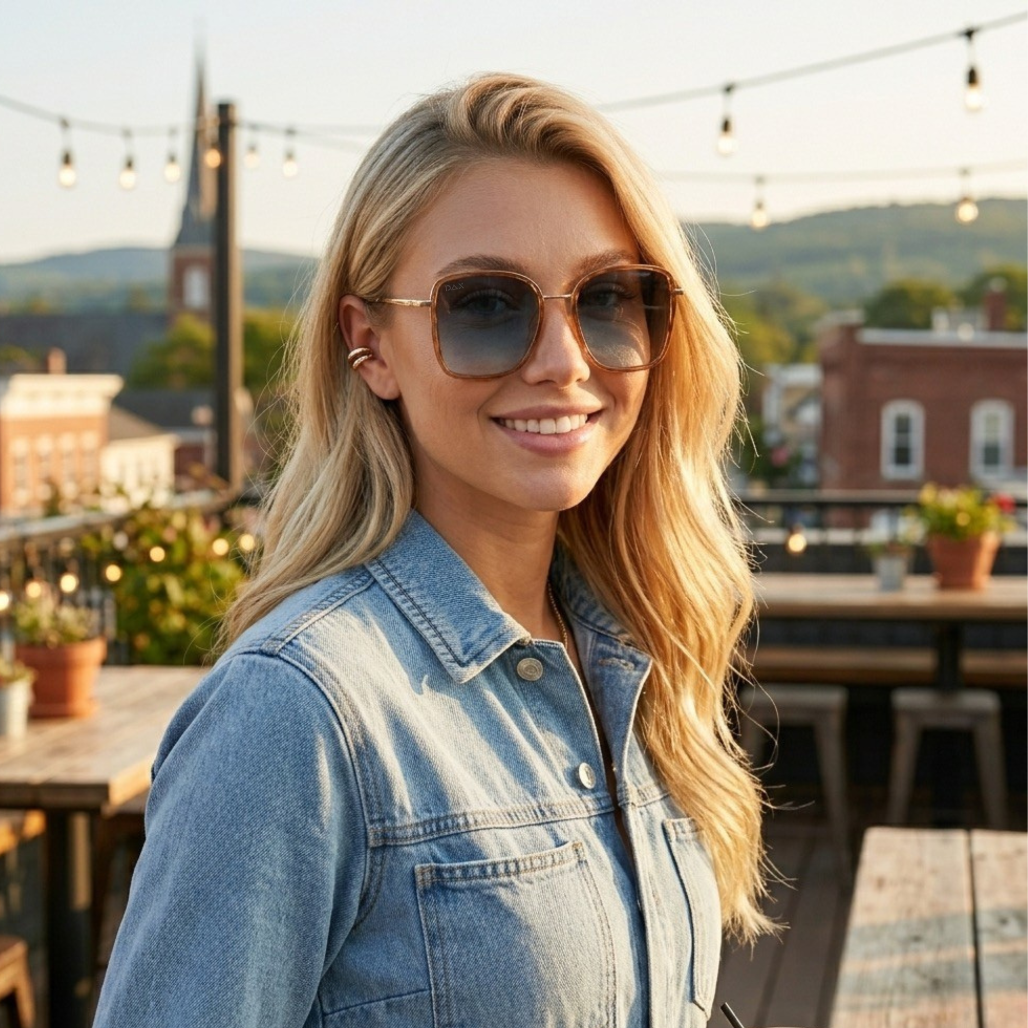 Woman wearing sunglasses and a denim jacket on a rooftop with a scenic background