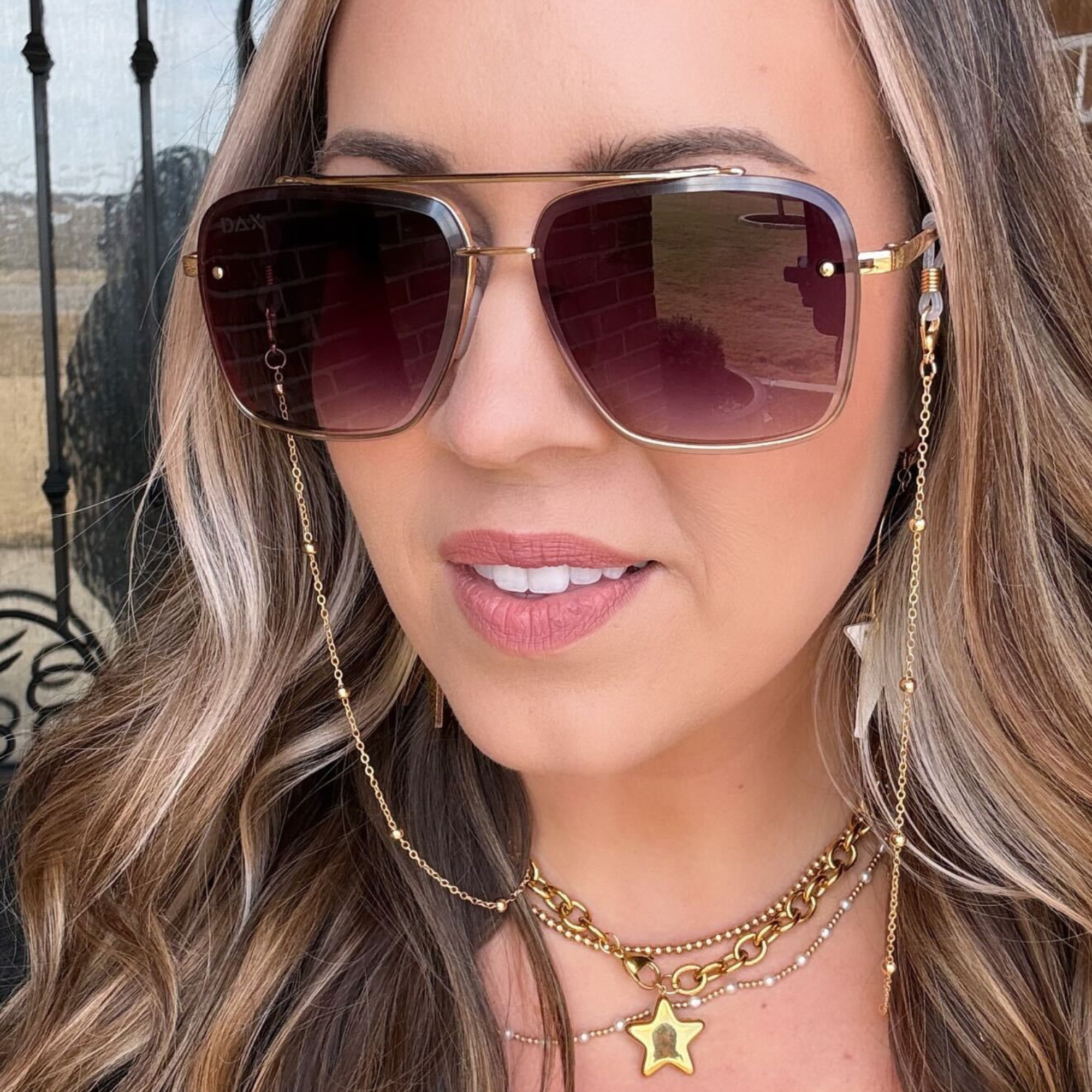 Woman wearing sunglasses with a gold chain, smiling outdoors.