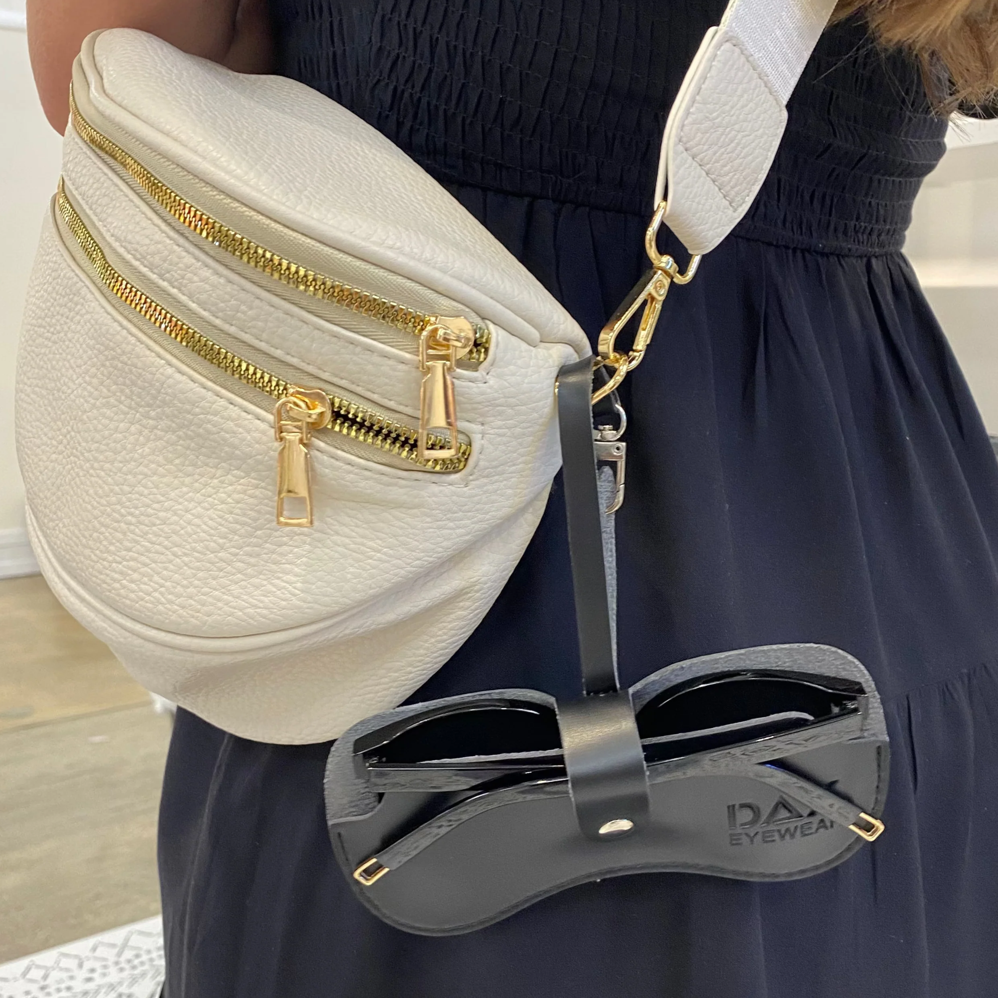 White waist bag with gold zippers held by a person wearing a navy dress, with sunglasses hanging from it.