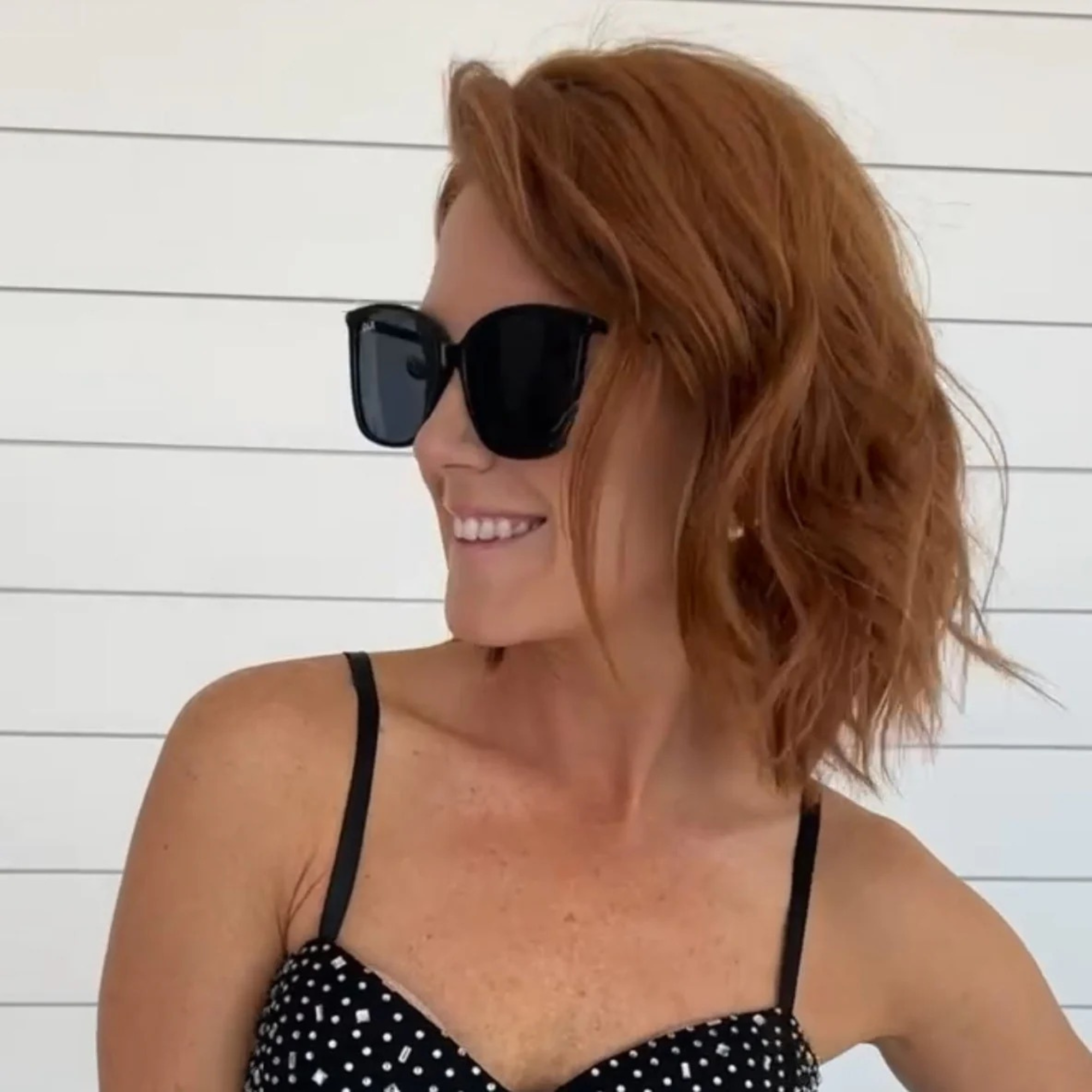 Woman wearing sunglasses and a black polka dot top against a white wooden background