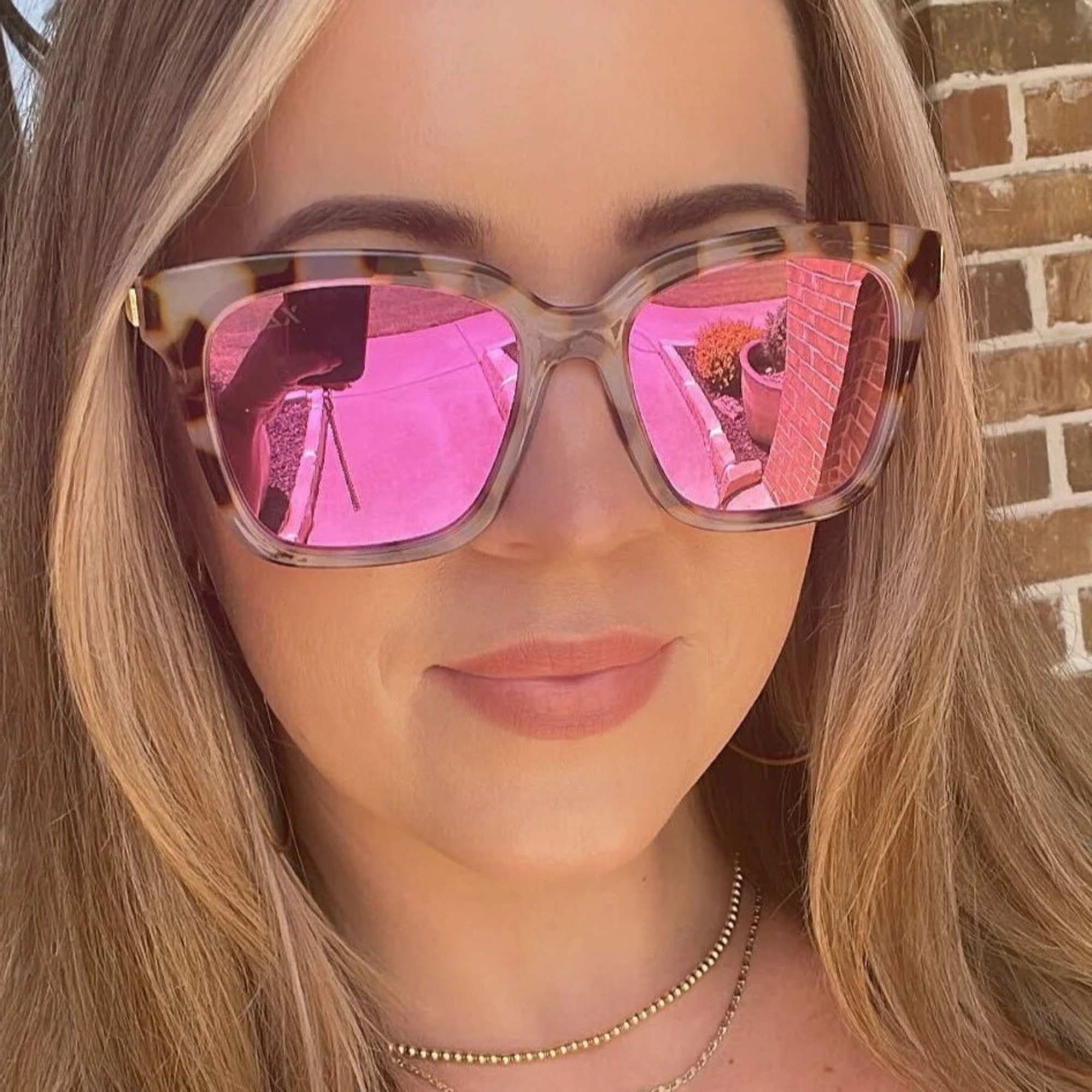 Person wearing sunglasses with pink reflection, standing against a brick wall.