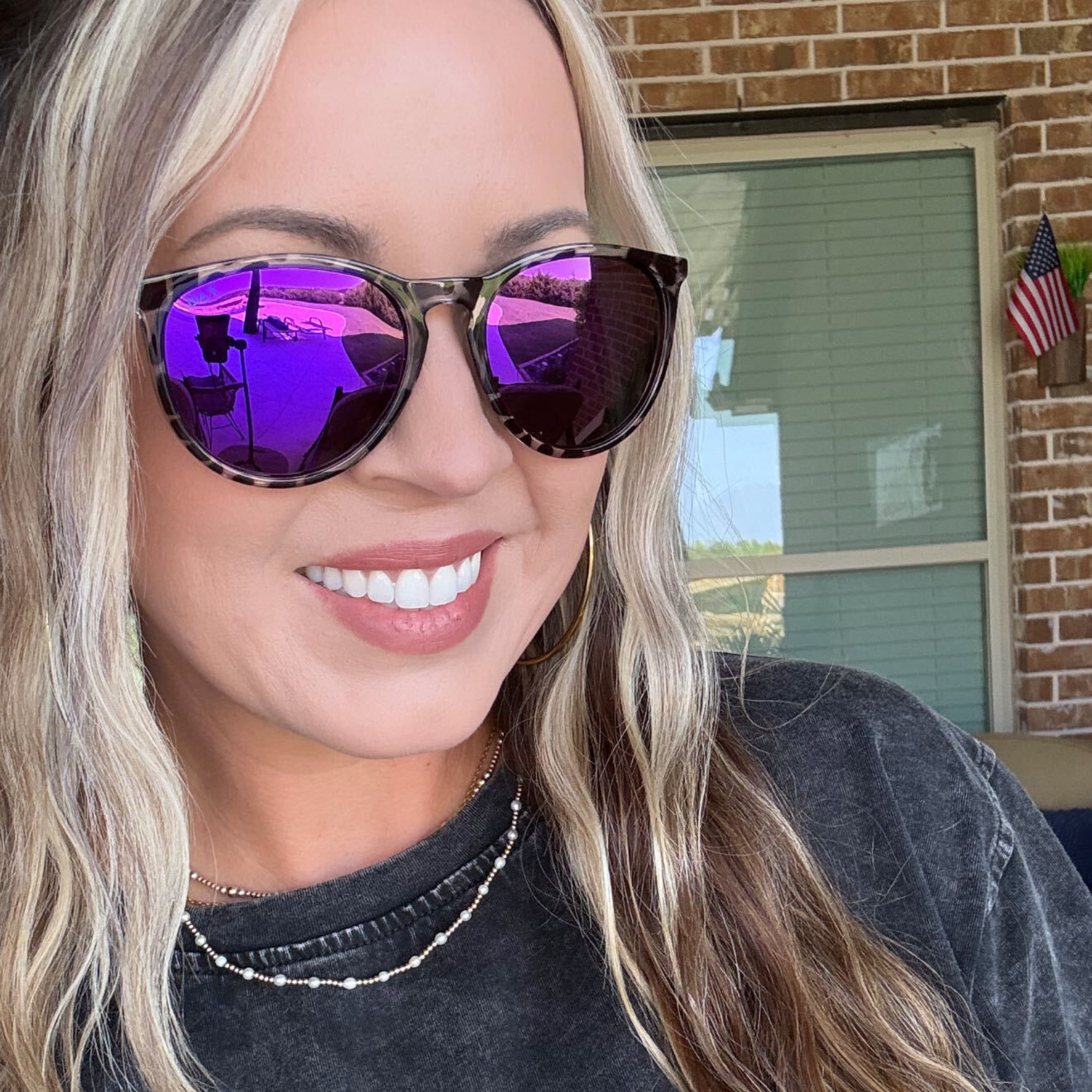 A close-up lifestyle shot of a woman with blonde hair wearing tortoiseshell sunglasses with vibrant purple mirrored lenses and a black t-shirt, set against a brick wall background.