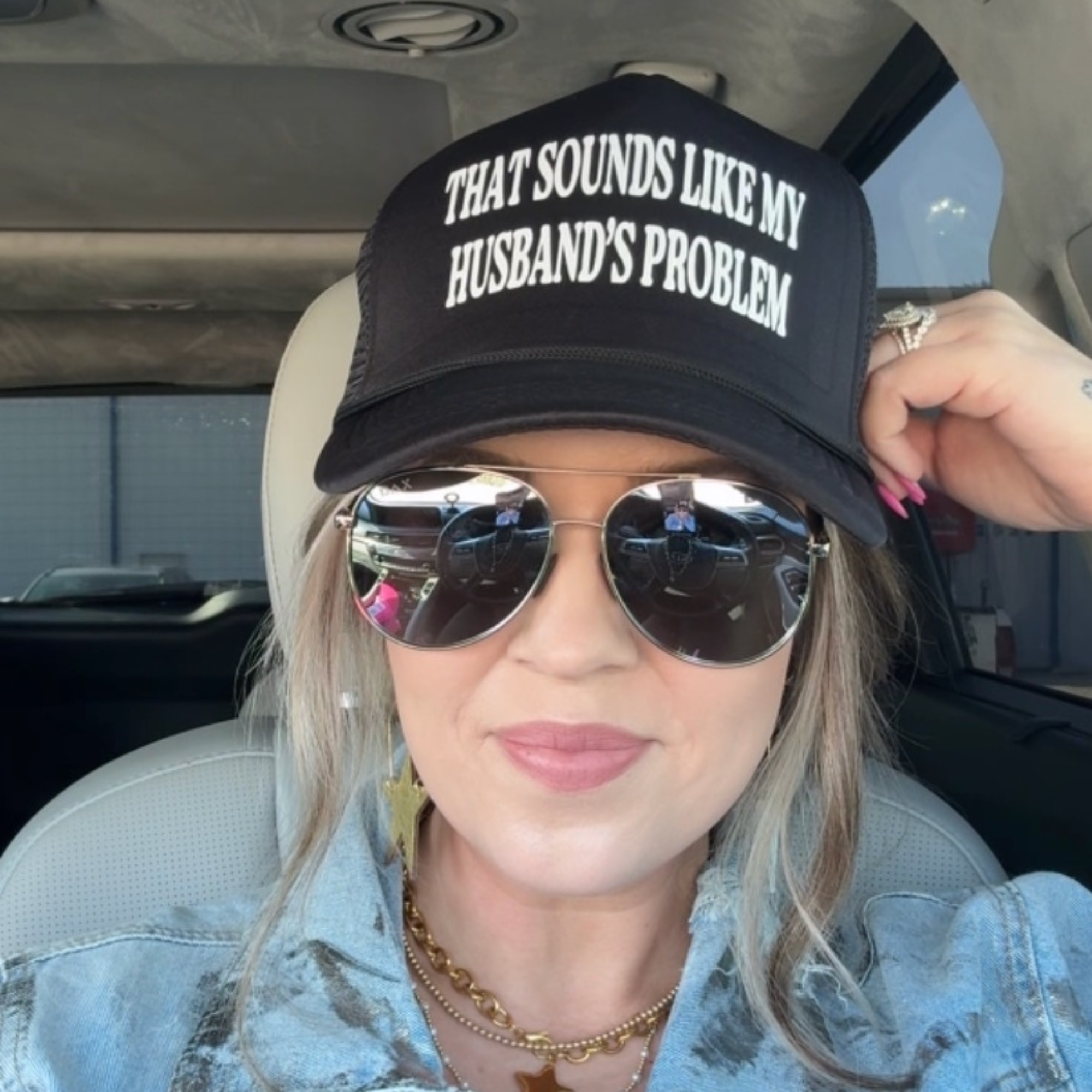 Person wearing a black cap with text and sunglasses, sitting inside a vehicle.