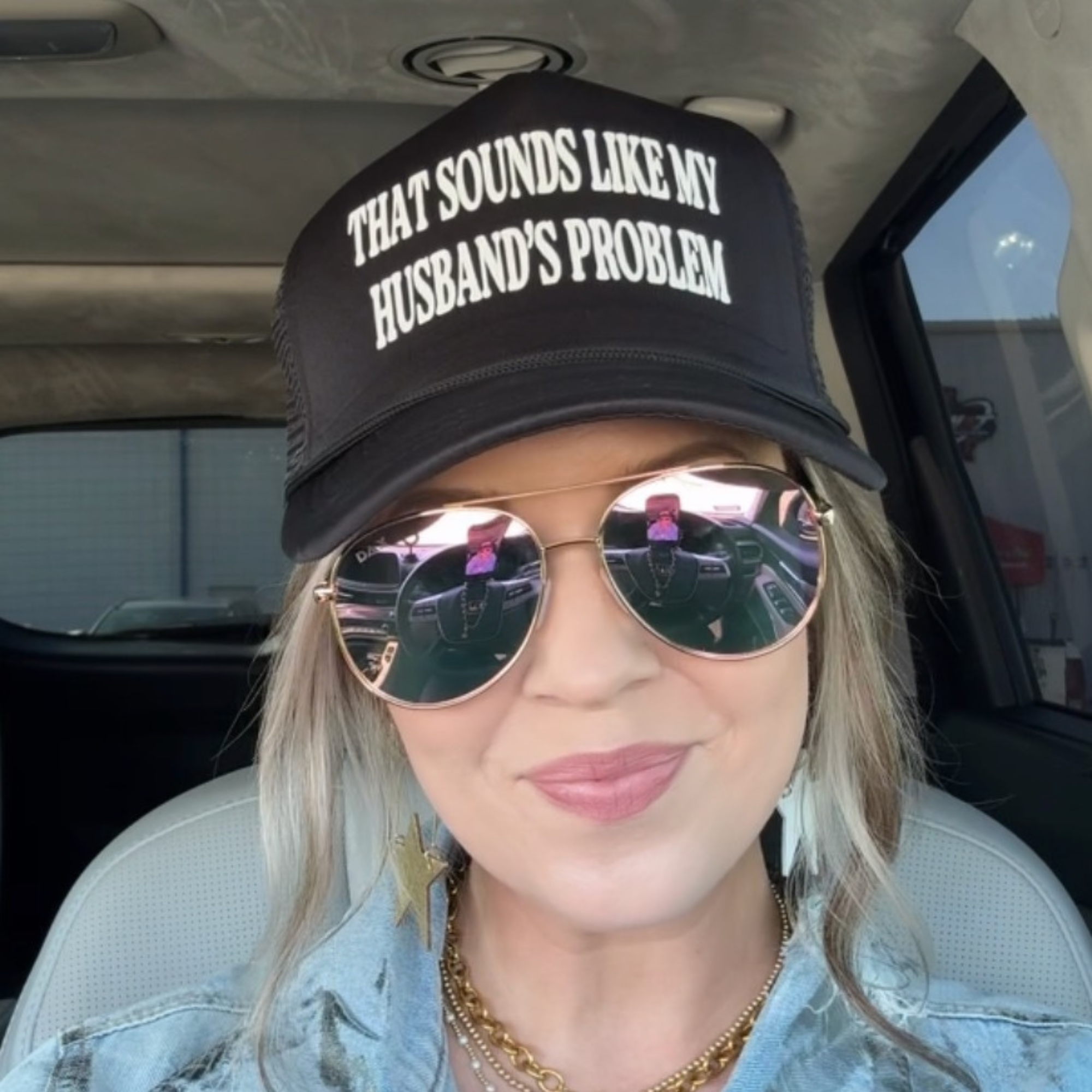 Person wearing a black cap with text and sunglasses, sitting inside a vehicle.