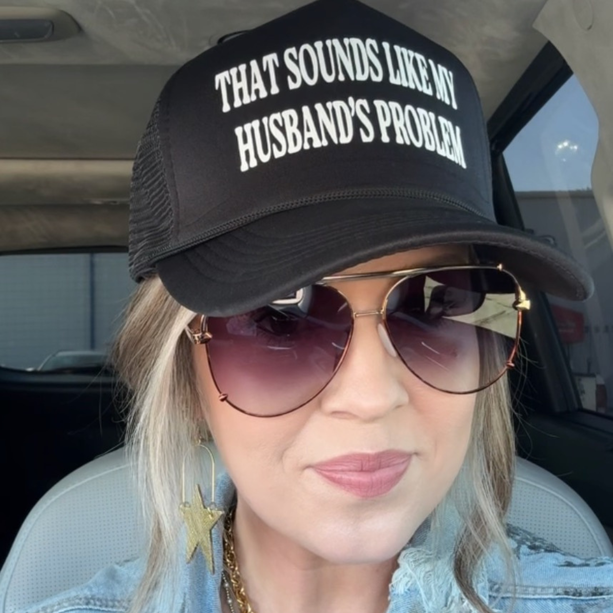 Person wearing a black cap with text and sunglasses, sitting inside a vehicle.