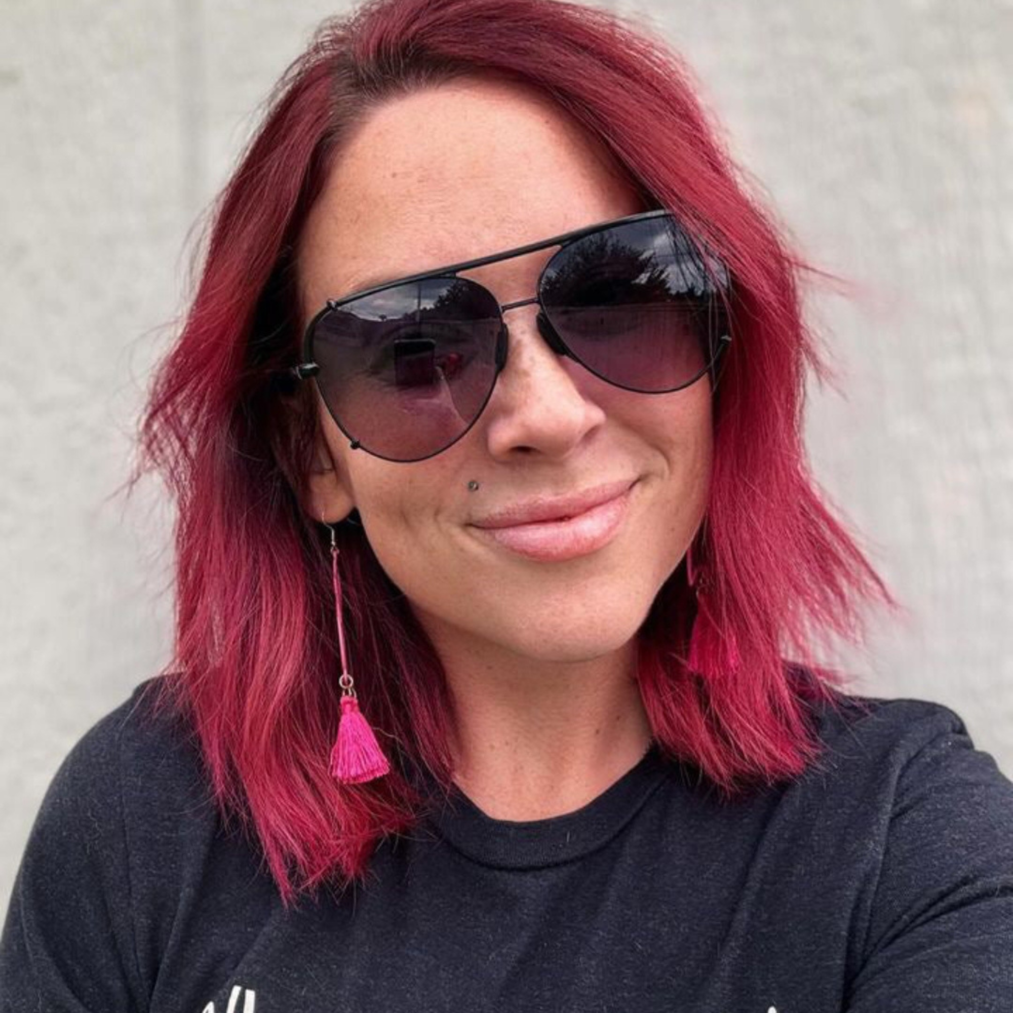 Person with pink hair wearing sunglasses and a black shirt against a neutral background