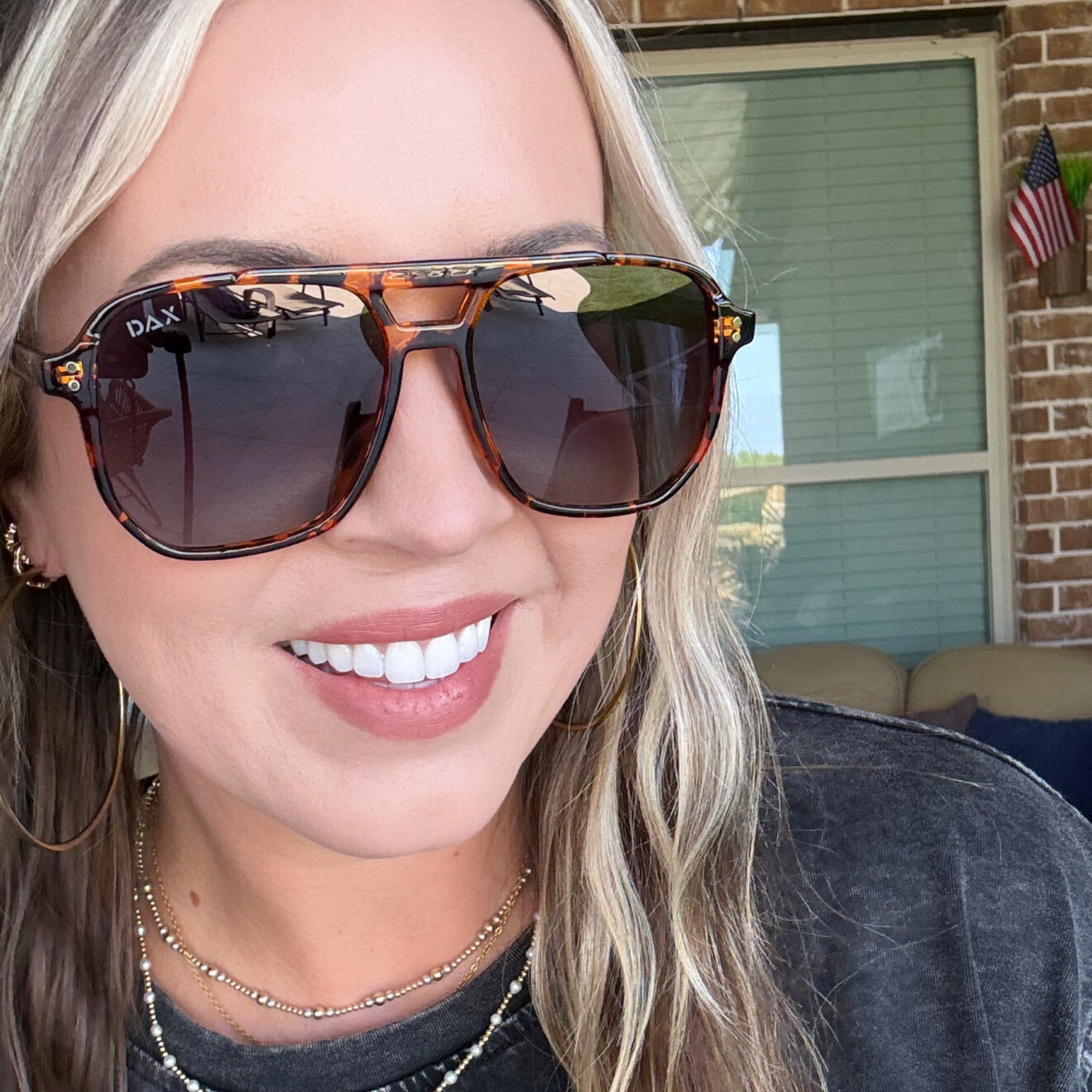 A close-up lifestyle shot of a woman wearing DAX brand aviator-style sunglasses with a brown tortoiseshell frame and dark lenses, set against a residential porch background.