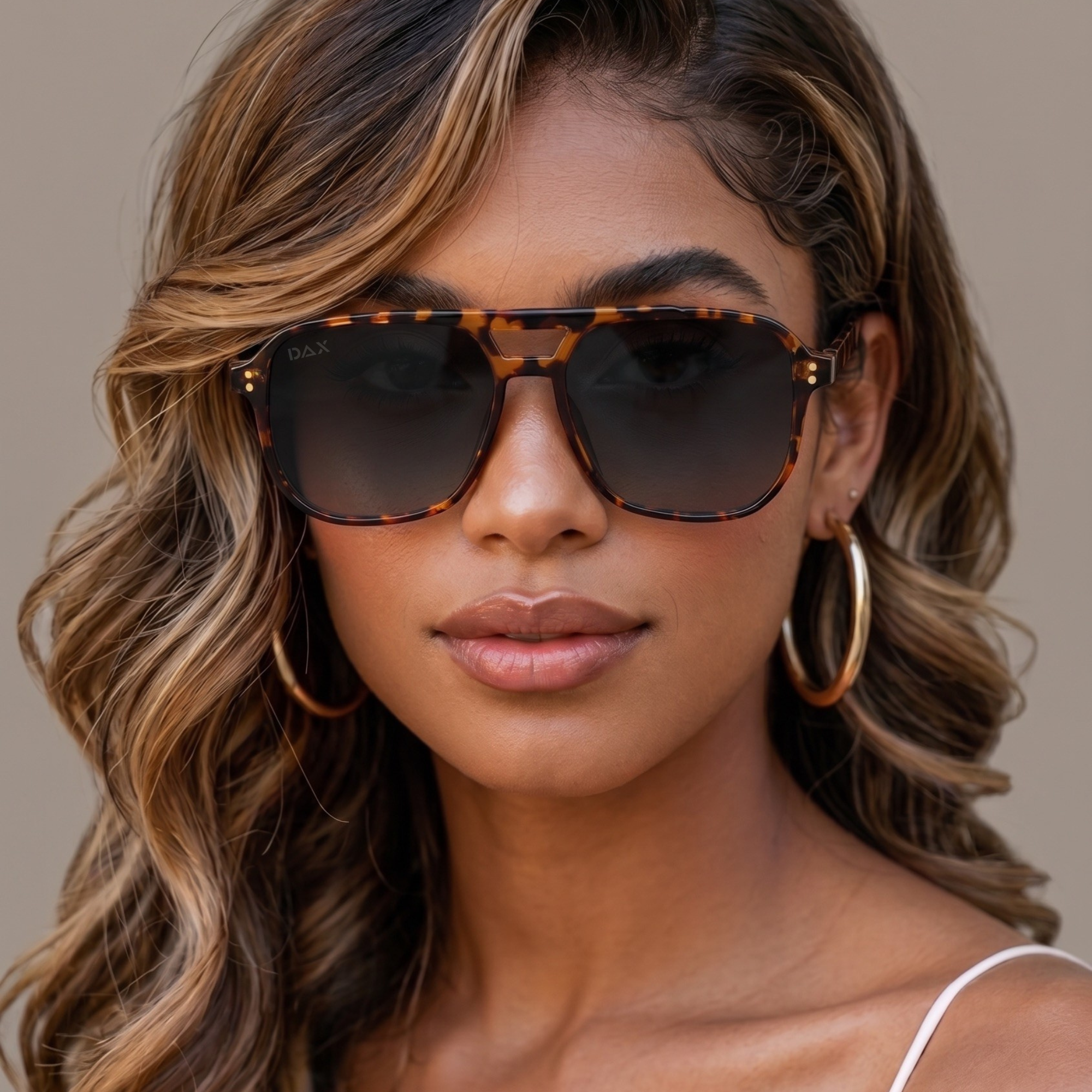 A close-up of a woman wearing aviator-style sunglasses with a tortoiseshell pattern and dark lenses, paired with gold hoop earrings against a neutral beige background.