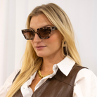 Woman wearing tortoiseshell sunglasses, a brown vest, and a white shirt against a plain background.