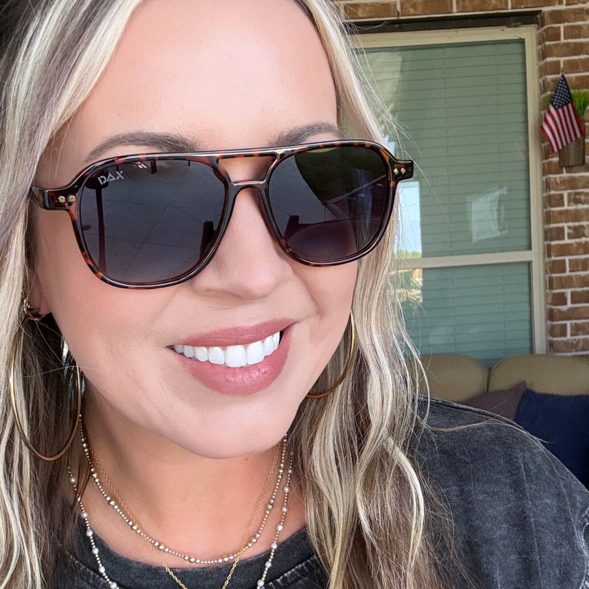 A close-up of a smiling woman wearing DAK sunglasses with dark lenses and tortoiseshell-style frames, set against a home porch background.