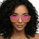 Woman wearing gold-framed sunglasses with pink lenses on a neutral background