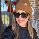Person wearing a brown beanie with a logo and dark sunglasses, standing in front of a brick wall.