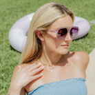 A blonde woman wearing light purple sunglasses while sitting in the grass.