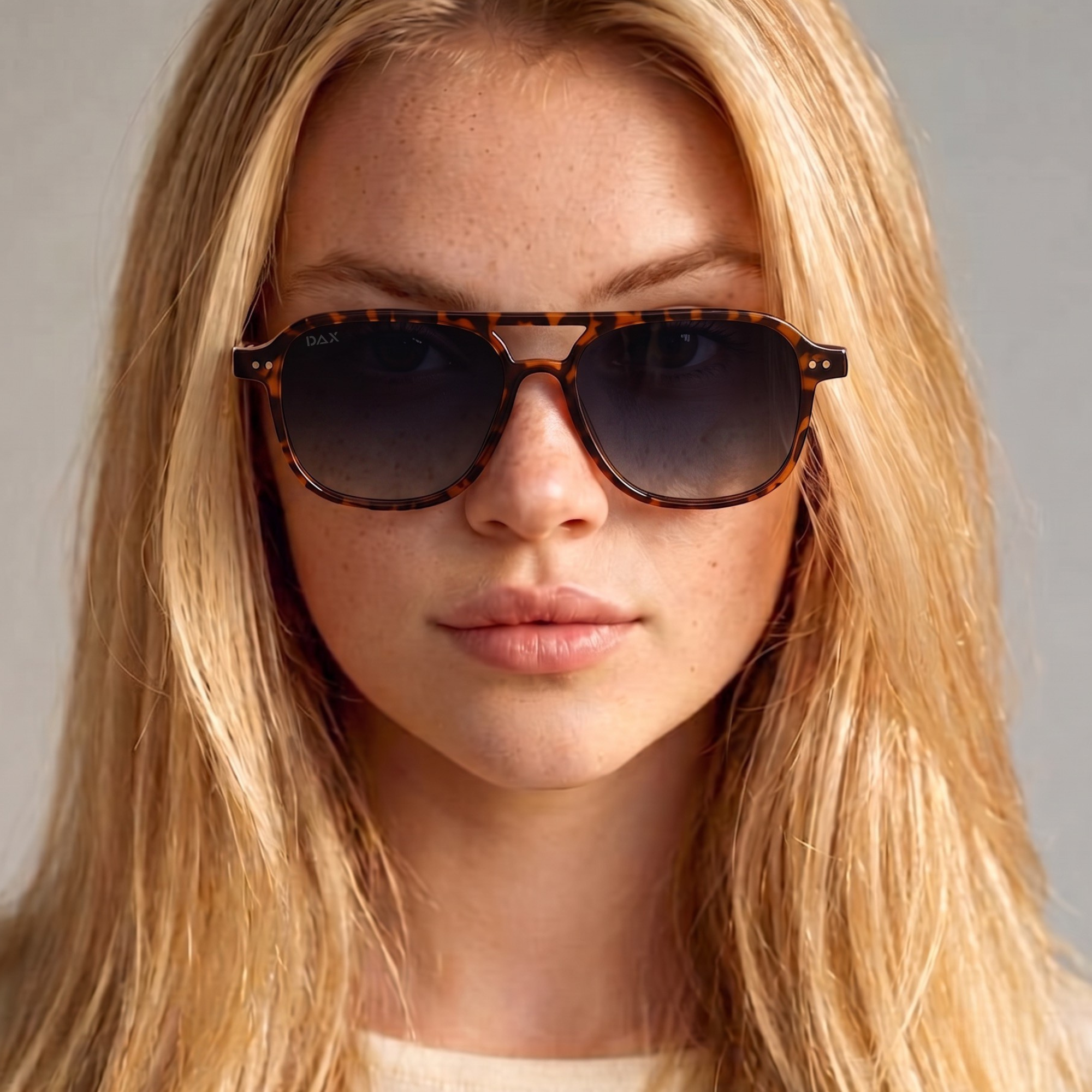 A close-up of a woman with blonde hair wearing tortoiseshell sunglasses with dark gradient lenses, set against a neutral gray background.