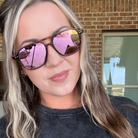A close-up of a woman wearing tortoiseshell sunglasses with pink mirrored lenses, photographed outdoors in front of a brick building.