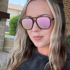 A close-up shot of a woman wearing tortoiseshell sunglasses with pink mirrored lenses, set against an outdoor residential background.
