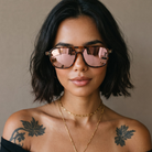 A close-up of a woman with short dark hair wearing oversized square sunglasses with reflective pink lenses and tortoiseshell-style frames, against a plain beige background.