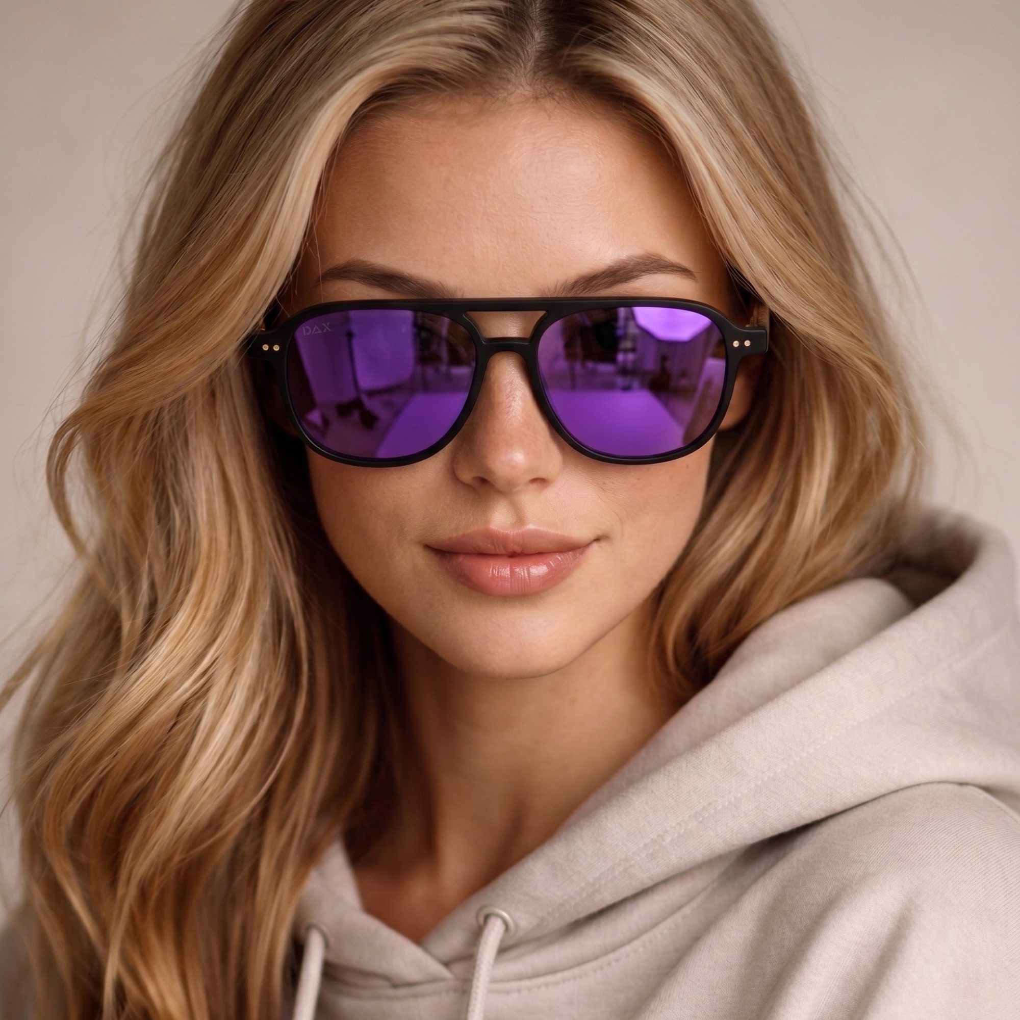 Woman wearing a light gray hoodie and purple-lensed sunglasses against a neutral background