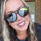 A close-up lifestyle shot of a woman wearing black aviator-style sunglasses with dark mirrored lenses, set against a background of a brick wall and window.