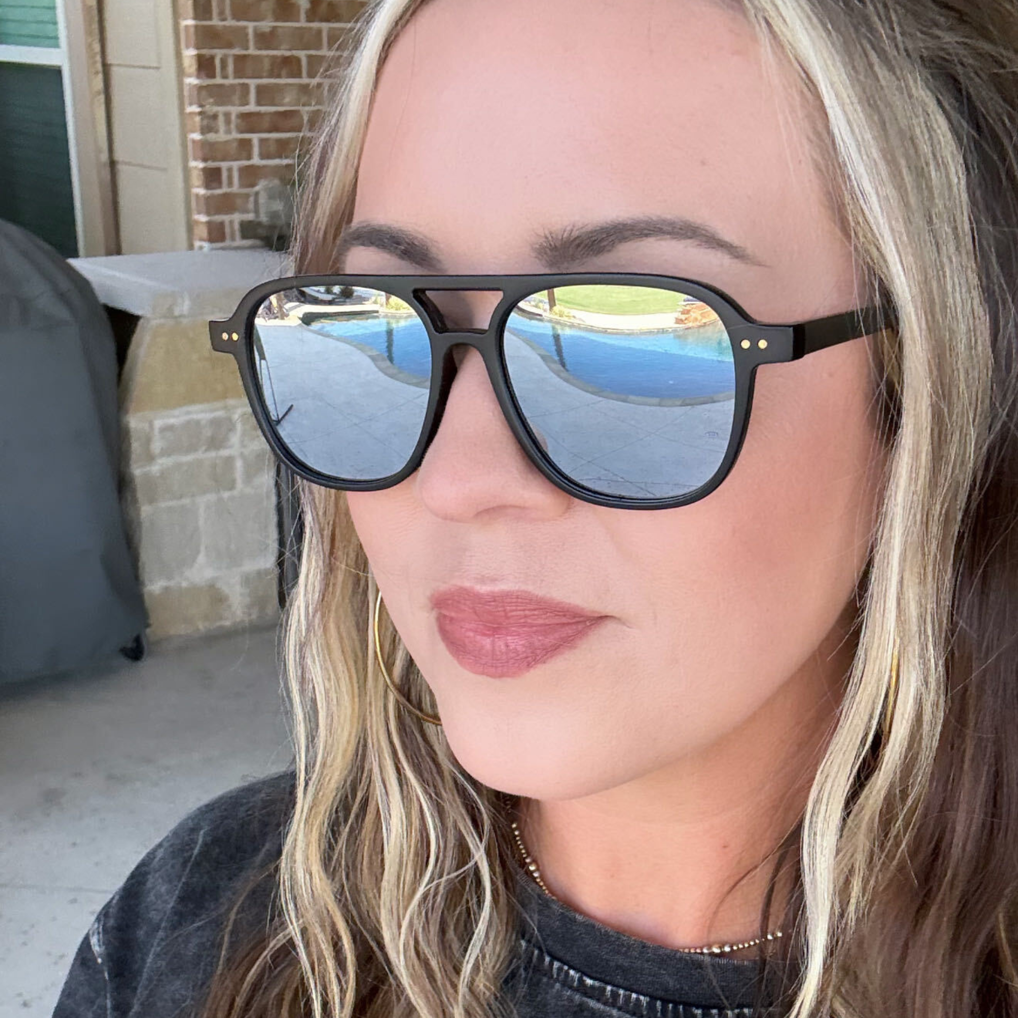 A close-up of a woman wearing black square sunglasses with mirrored blue lenses, photographed outdoors in front of a brick building.