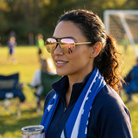 Woman wearing sunglasses and a blue scarf, standing on a grassy field with a soccer goal in the background.