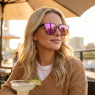Woman wearing sunglasses with a cityscape reflection, holding a margarita outdoors.