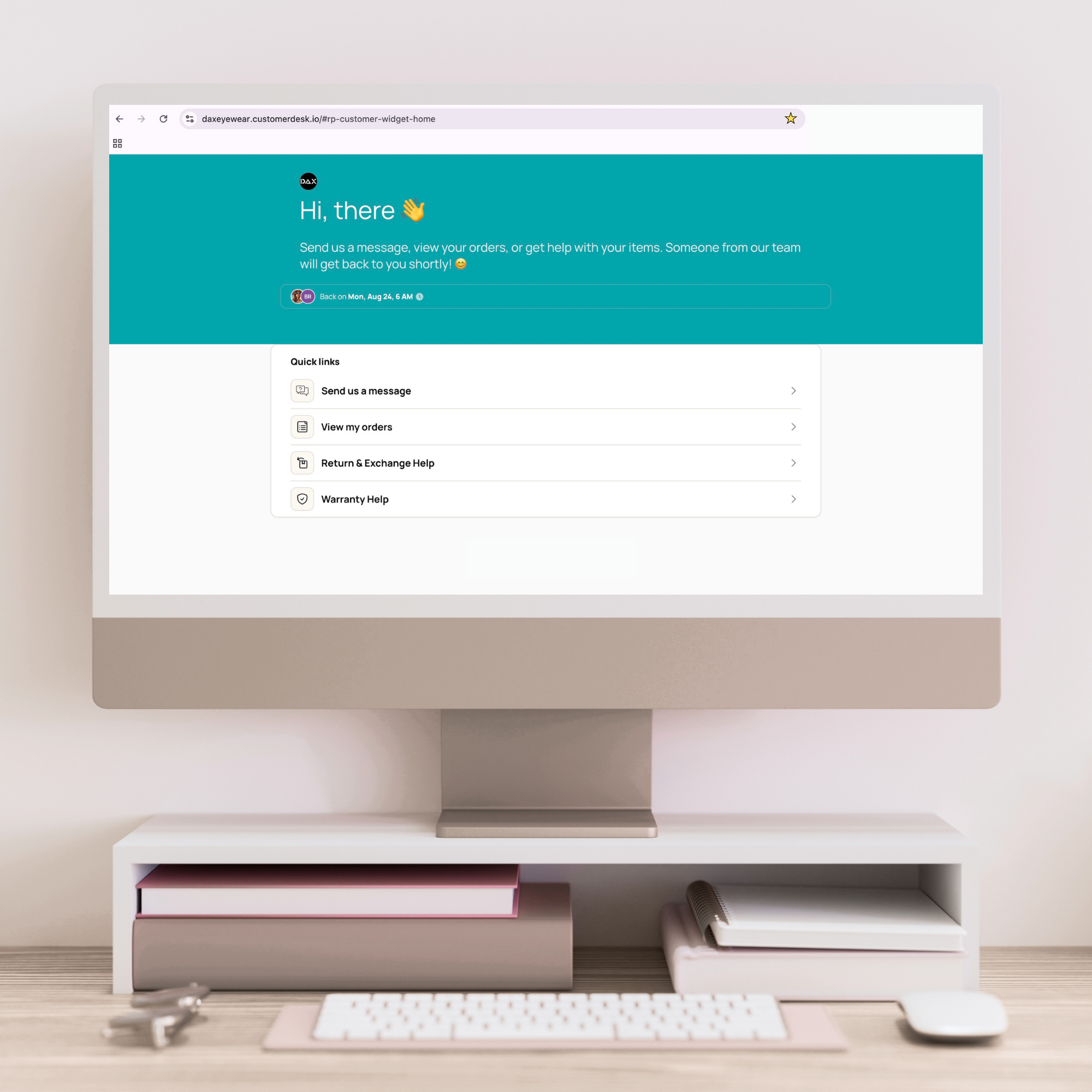 A desktop computer monitor on a beige monitor stand, with a keyboard and mouse on a light wood desk. The screen displays a customer service chat interface with a teal and white design.