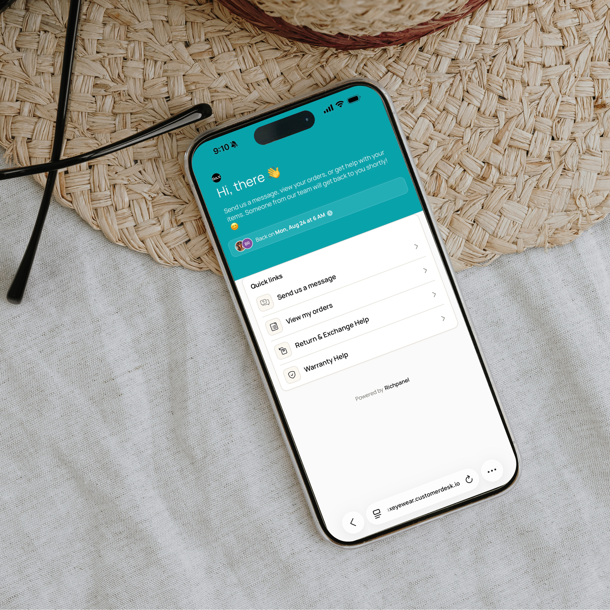 A smartphone lying on a light fabric surface beside a woven straw mat, with black eyeglasses visible. The phone screen shows a teal and white messaging app interface.