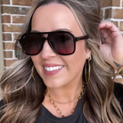 Woman wearing sunglasses and gold jewelry against a brick wall.
