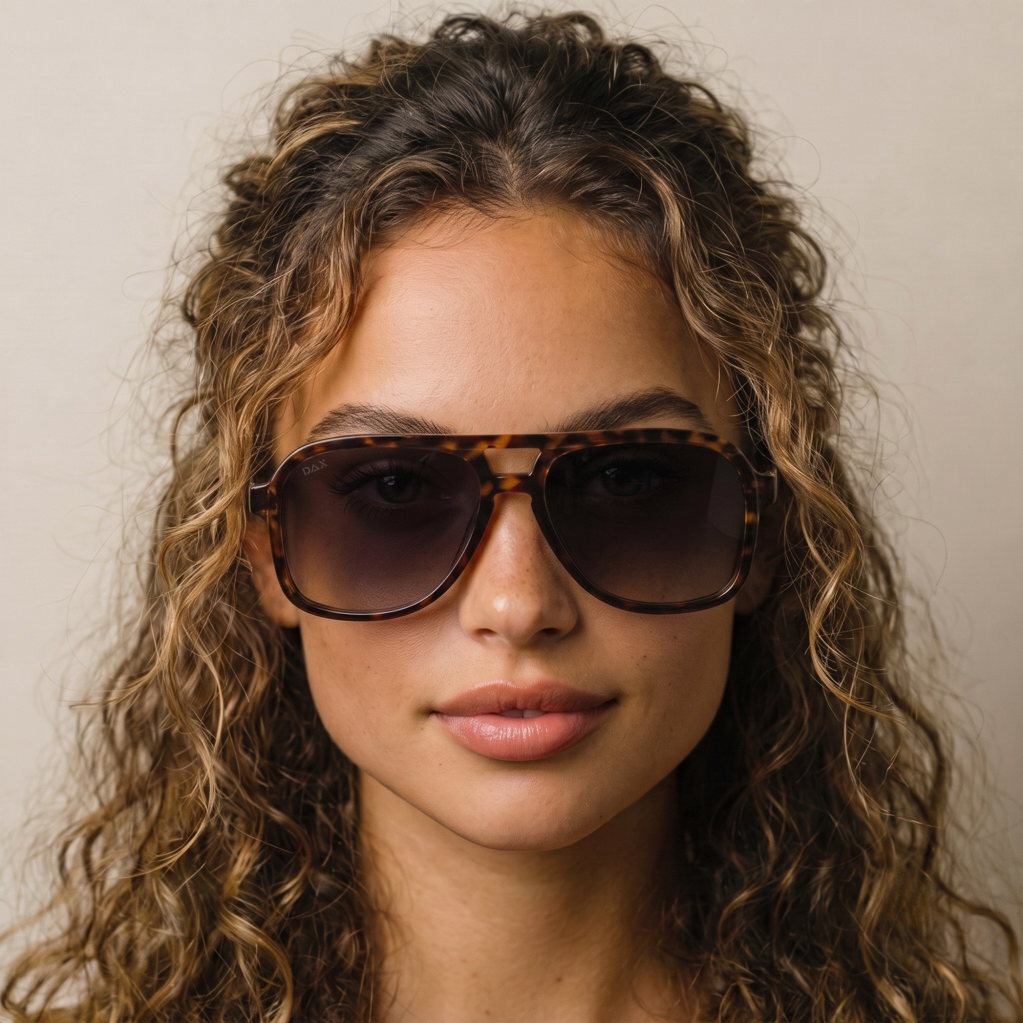 A close-up portrait of a woman with curly brown hair wearing oversized square sunglasses with a tortoiseshell frame and dark tinted lenses, set against a plain light beige background.