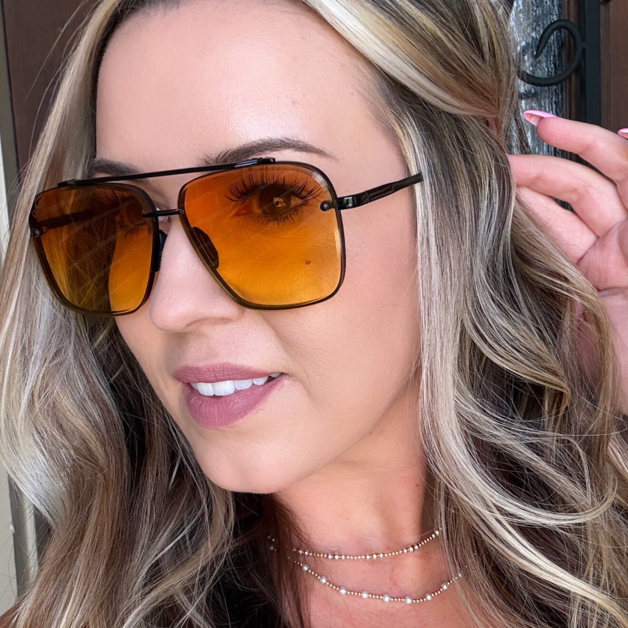 A close-up of a woman with blonde hair wearing square aviator-style sunglasses with amber-orange tinted lenses and black metal frames.