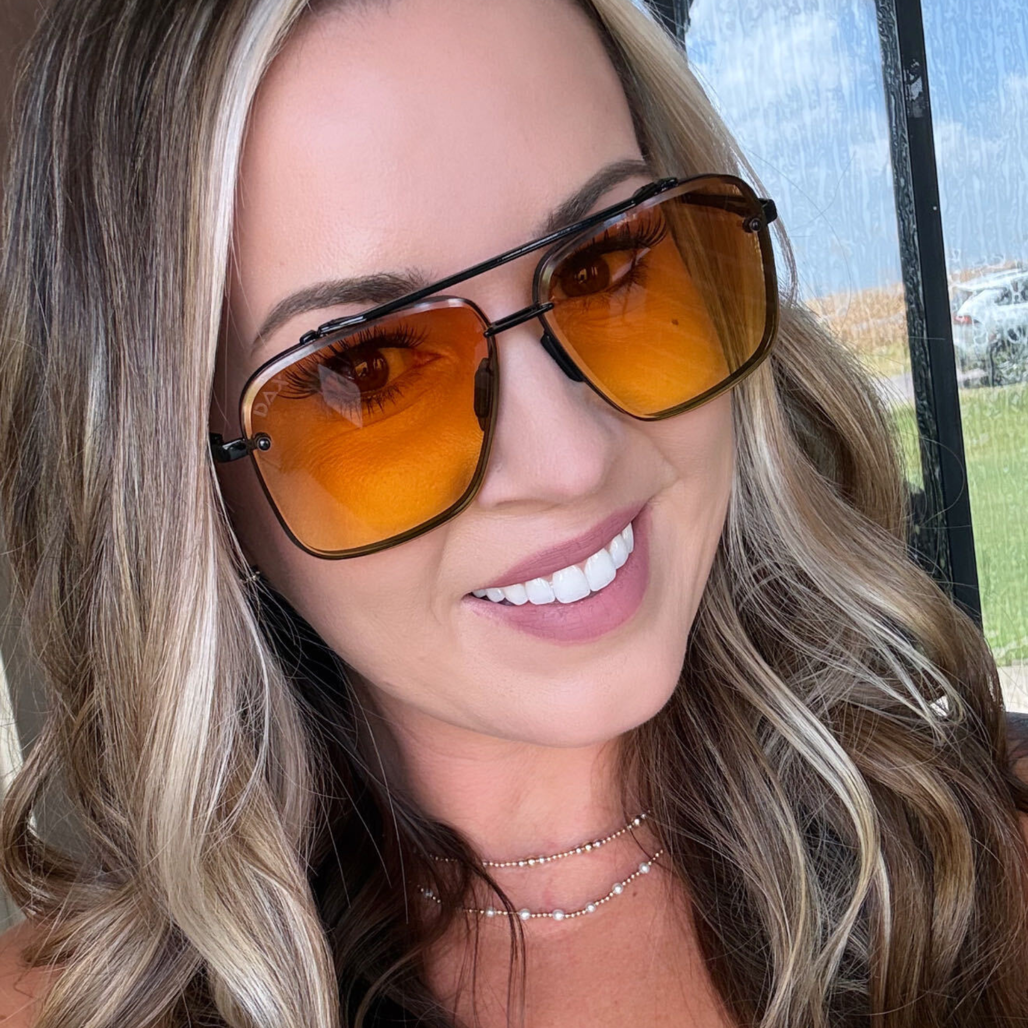 A close-up of a smiling woman with blonde hair wearing square aviator-style sunglasses with orange-tinted lenses and a thin black frame.