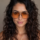 A close-up portrait of a woman with long, dark, curly hair wearing aviator-style sunglasses with amber-orange tinted lenses and thin dark metal frames.