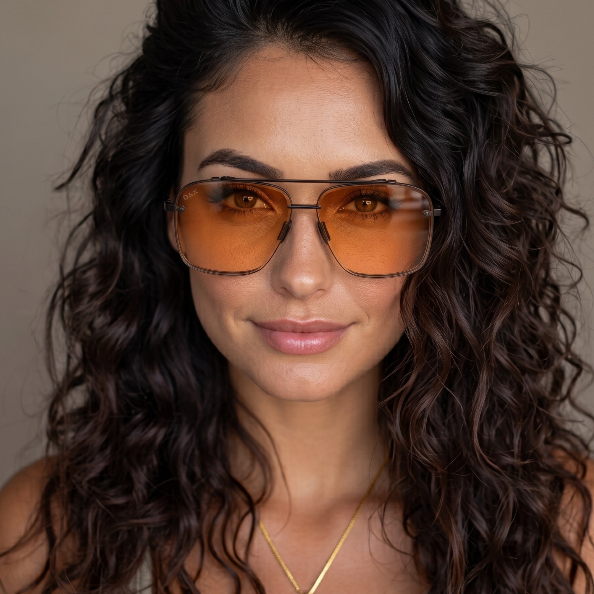 A close-up portrait of a woman with long, dark, curly hair wearing aviator-style sunglasses with amber-orange tinted lenses and thin dark metal frames.