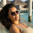 Woman wearing sunglasses and a beige top in a hammock by a waterfront.