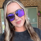 A woman wearing square aviator-style sunglasses with purple mirrored lenses and black frames, photographed outdoors in front of a brick building.