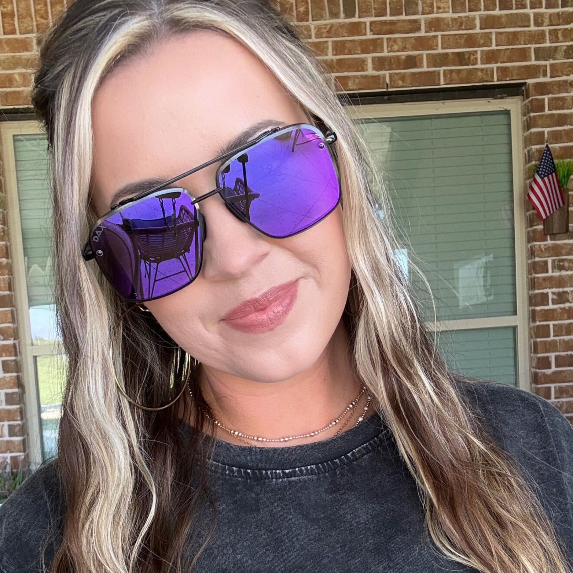 A woman wearing square aviator-style sunglasses with purple mirrored lenses and black frames, photographed outdoors in front of a brick building.