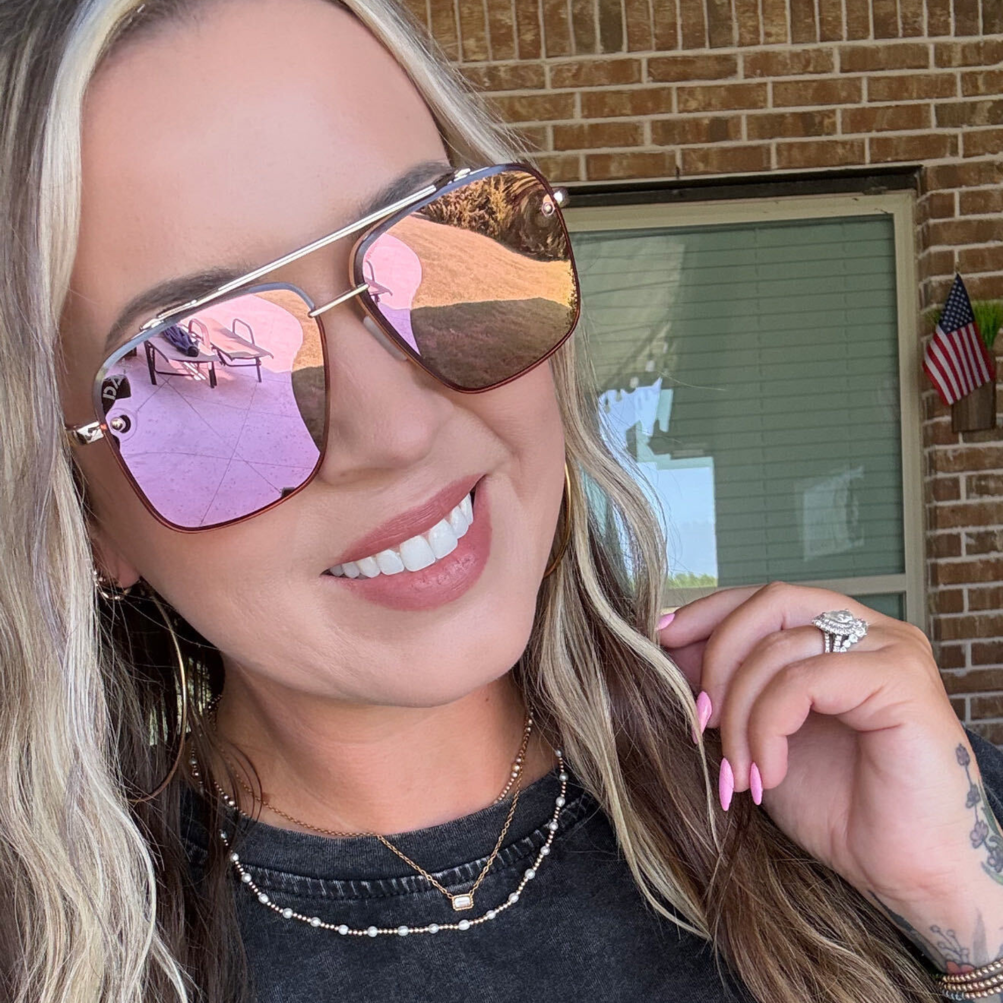 A close-up lifestyle shot of a woman wearing aviator sunglasses with reflective pink and gold mirrored lenses. She is smiling, wearing a black t-shirt and layered necklaces, with a brick wall and window in the background.