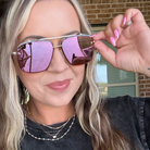 A close-up lifestyle shot of a woman wearing square aviator sunglasses with gold-toned metal frames and pink mirrored lenses, holding one lens with her hand. She is wearing a black t-shirt and layered gold necklaces, with a brick wall and window in the background.