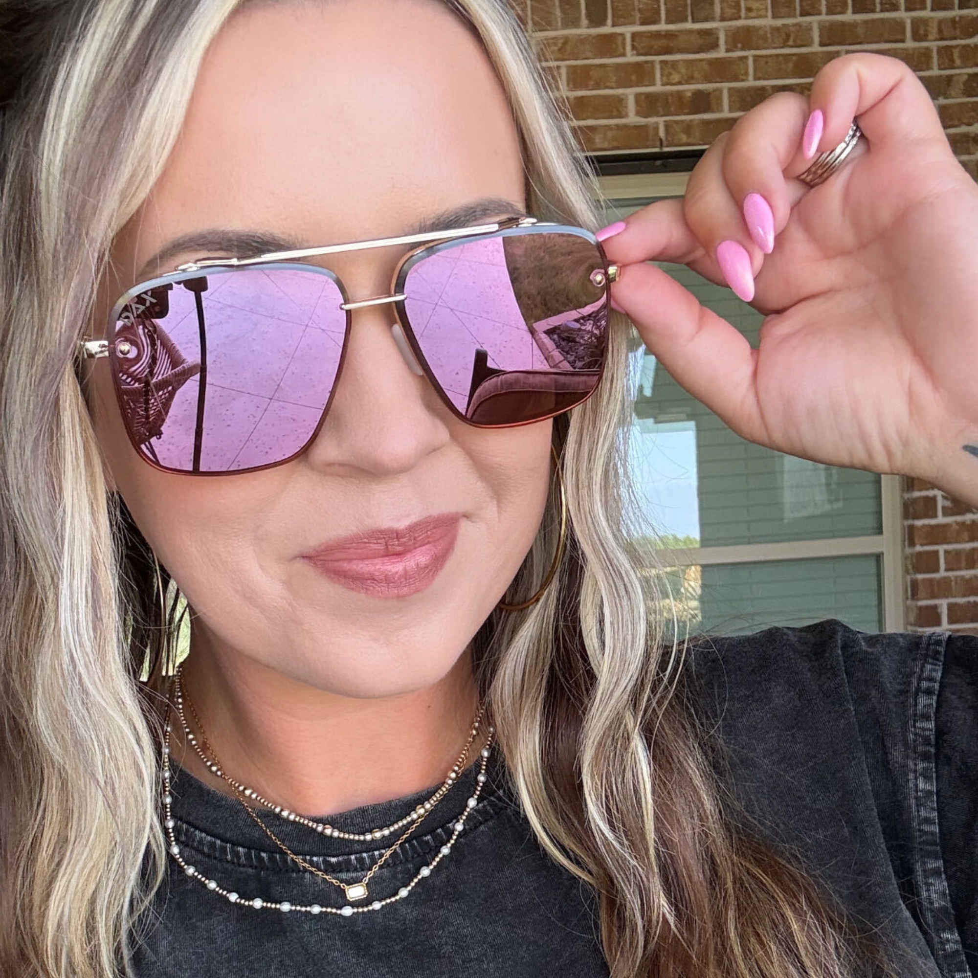 A close-up lifestyle shot of a woman wearing square aviator sunglasses with gold-toned metal frames and pink mirrored lenses, holding one lens with her hand. She is wearing a black t-shirt and layered gold necklaces, with a brick wall and window in the background.