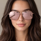 A close-up of a woman with long dark brown hair wearing oversized aviator-style sunglasses with a rose gold metal frame and pink-tinted lenses.