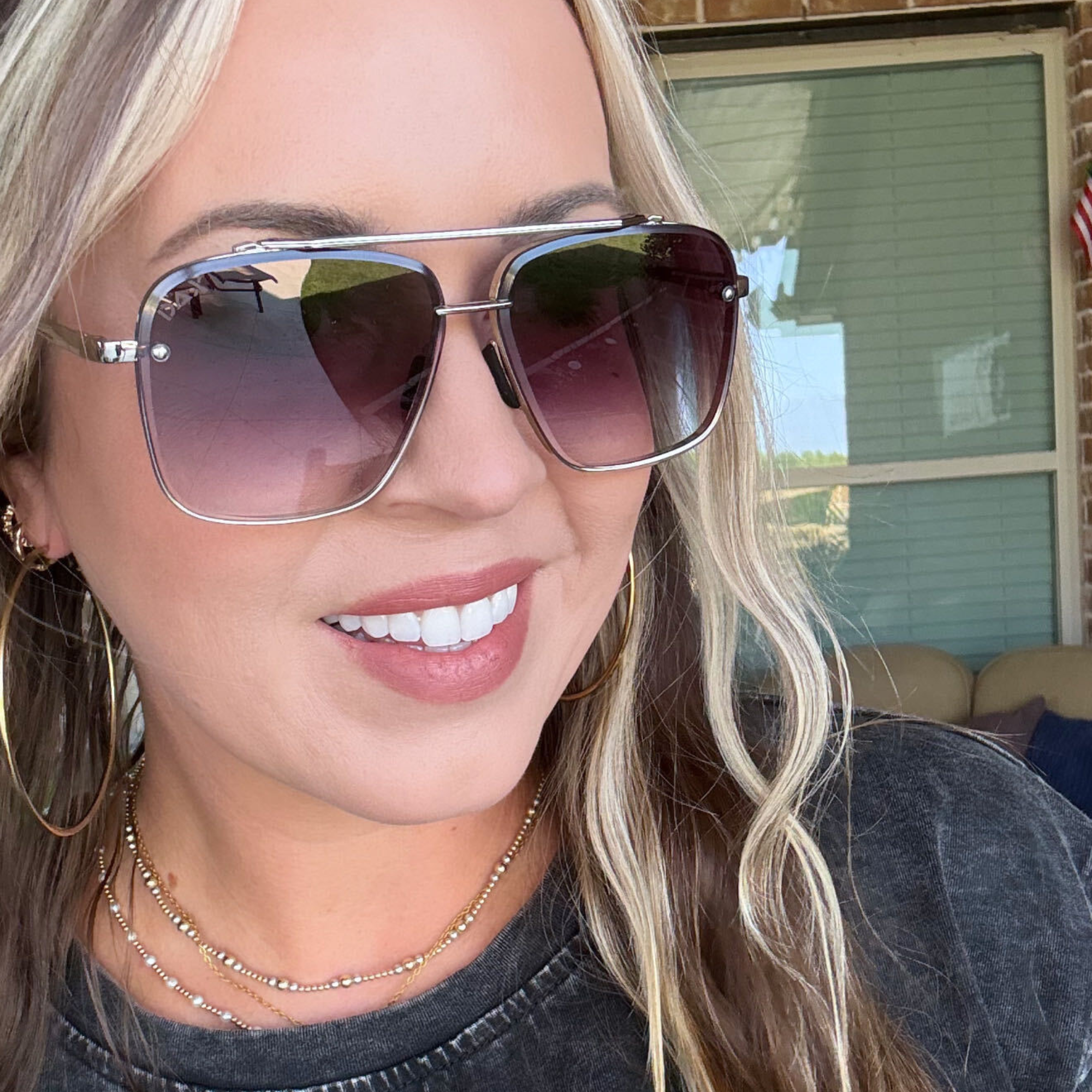 A close-up lifestyle shot of a woman wearing oversized square aviator sunglasses with a silver metal frame and gradient tinted lenses.