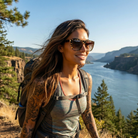 Woman with tattoos and sunglasses standing on a mountain overlooking a river and trees.