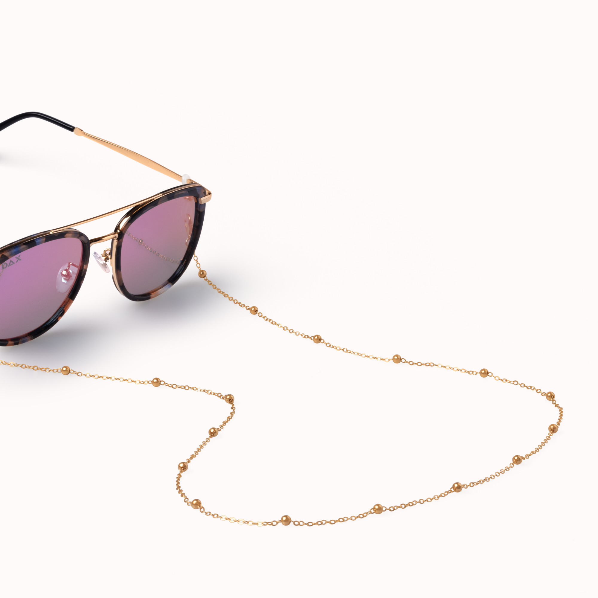 A gold chain sunglasses strap with small bead details, shown draped over the nose bridge of gold-framed sunglasses with pink-tinted lenses on a clean white background.