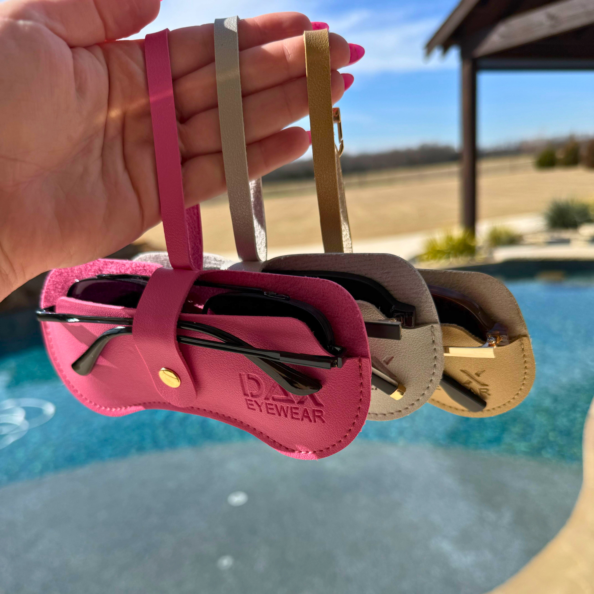 Three pairs of sunglasses in pink, silver, and gold cases held by a hand with a pool and outdoor setting in the background.